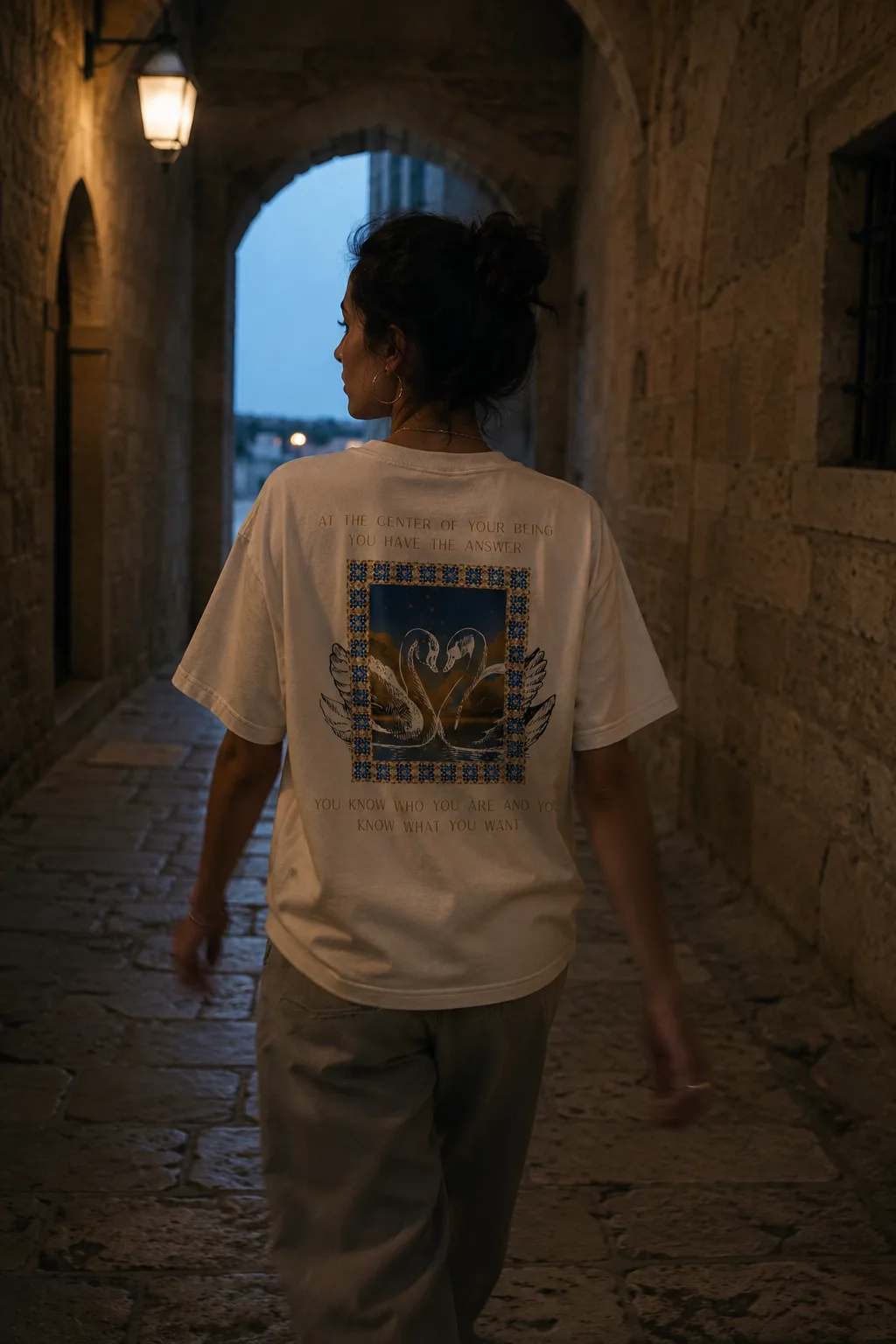 A woman walks through a lamplit stone archway at dusk wearing the Lao Tzu tee